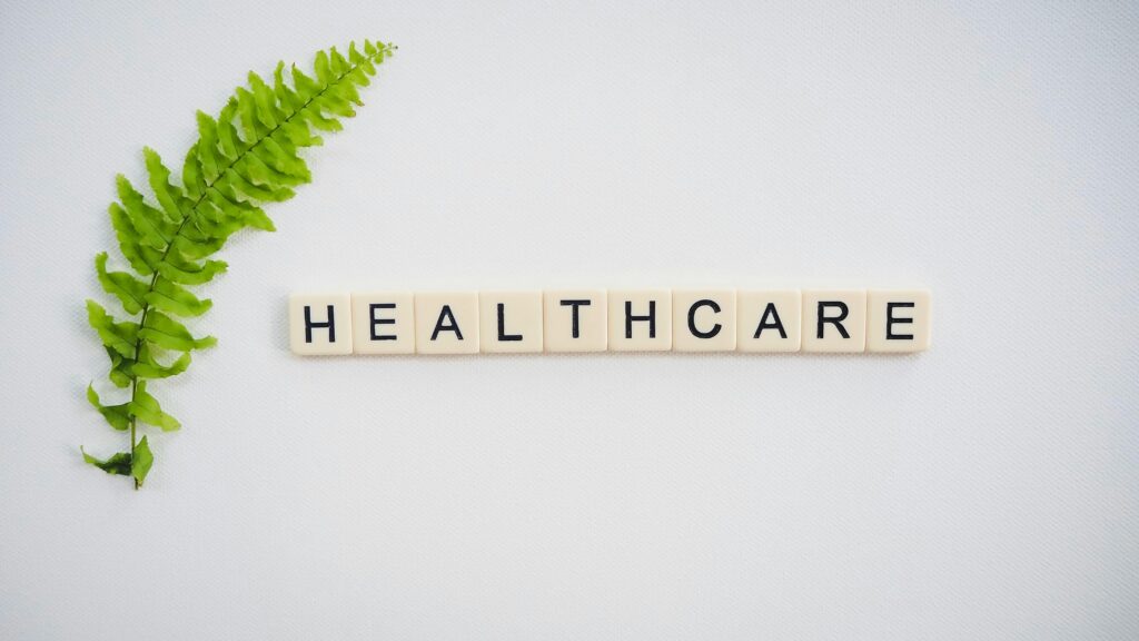 About Healthcare word spelled with Scrabble tiles next to a green fern leaf on a white background.